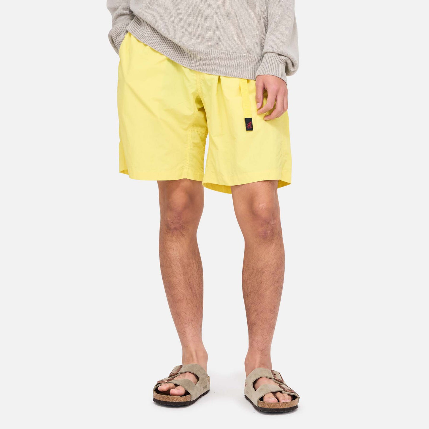 A person stands against a plain white background, wearing a gray sweater, Gramicci Nylon Loose Short in yellow (water-repellent, wind-resistant), and beige sandals.
