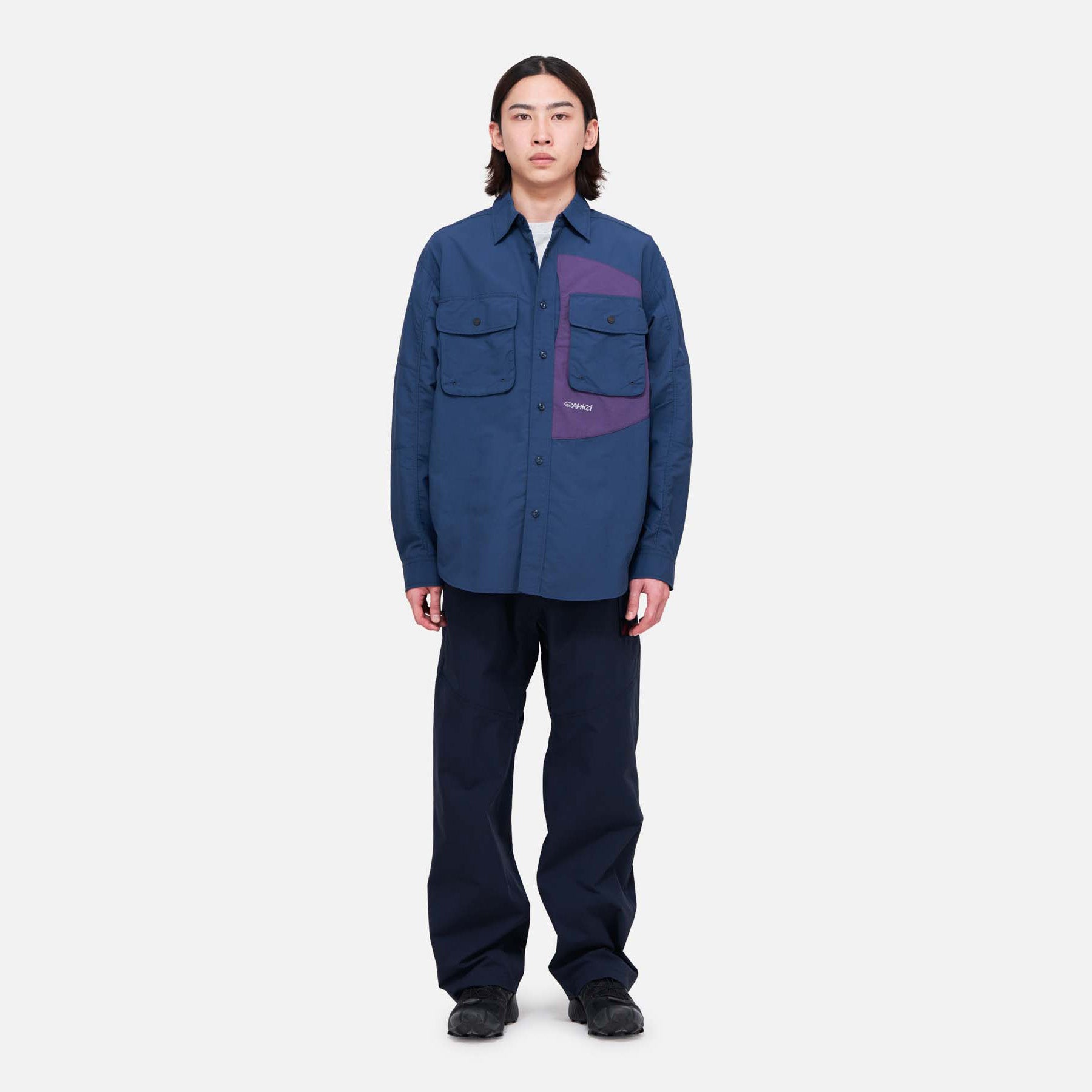 A person stands facing forward wearing Gramicci Trekking Pants with a blue button-up shirt and black shoes against a plain white background.