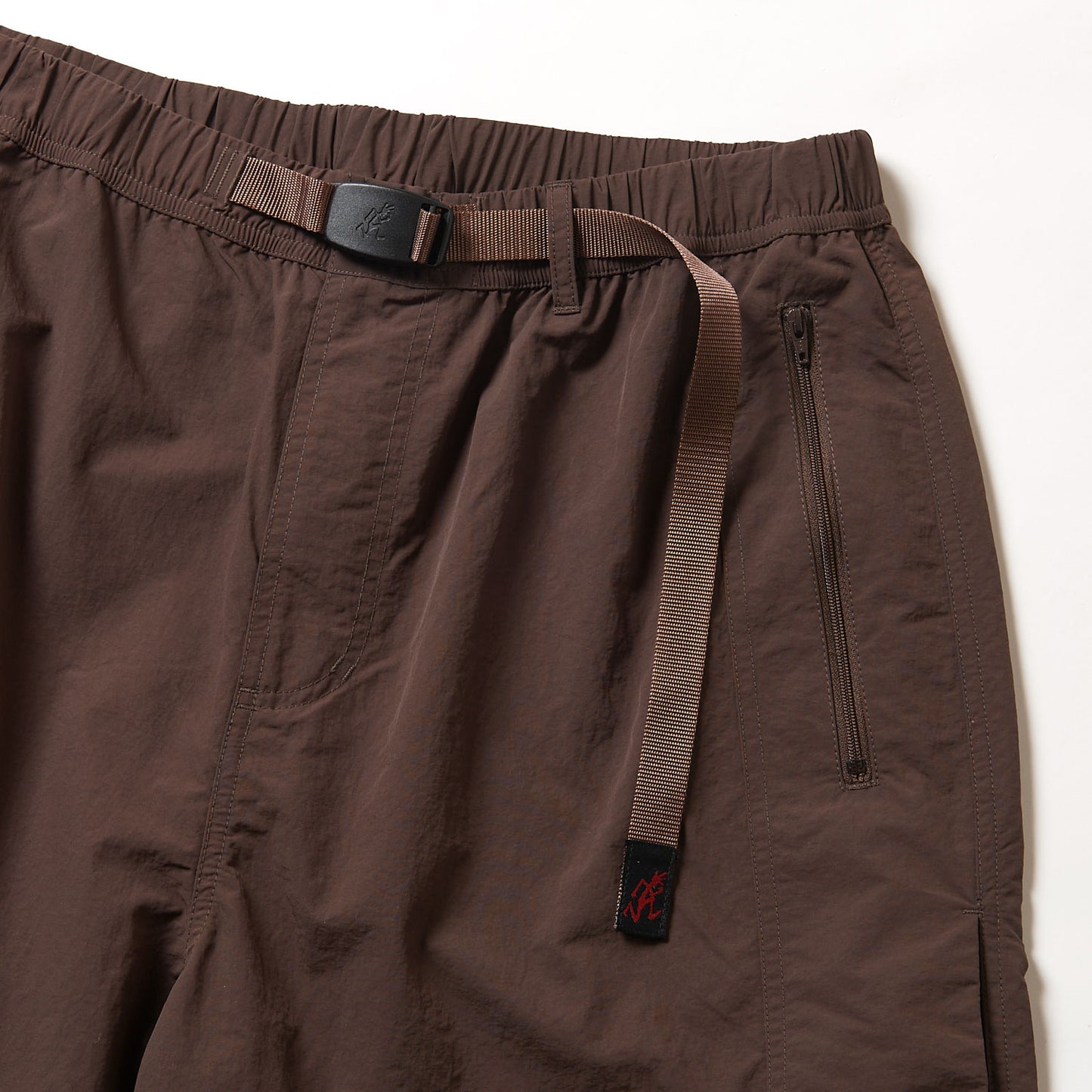 Close-up of Gramicci Nylon Runners in brown, showcasing an adjustable belt with a plastic buckle and a zippered side pocket, set against a white background.