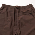 The Gramicci Nylon Runners are brown nylon pants with an elastic waistband, integrated belt, and a single back pocket accented by a small black and red woven tag.