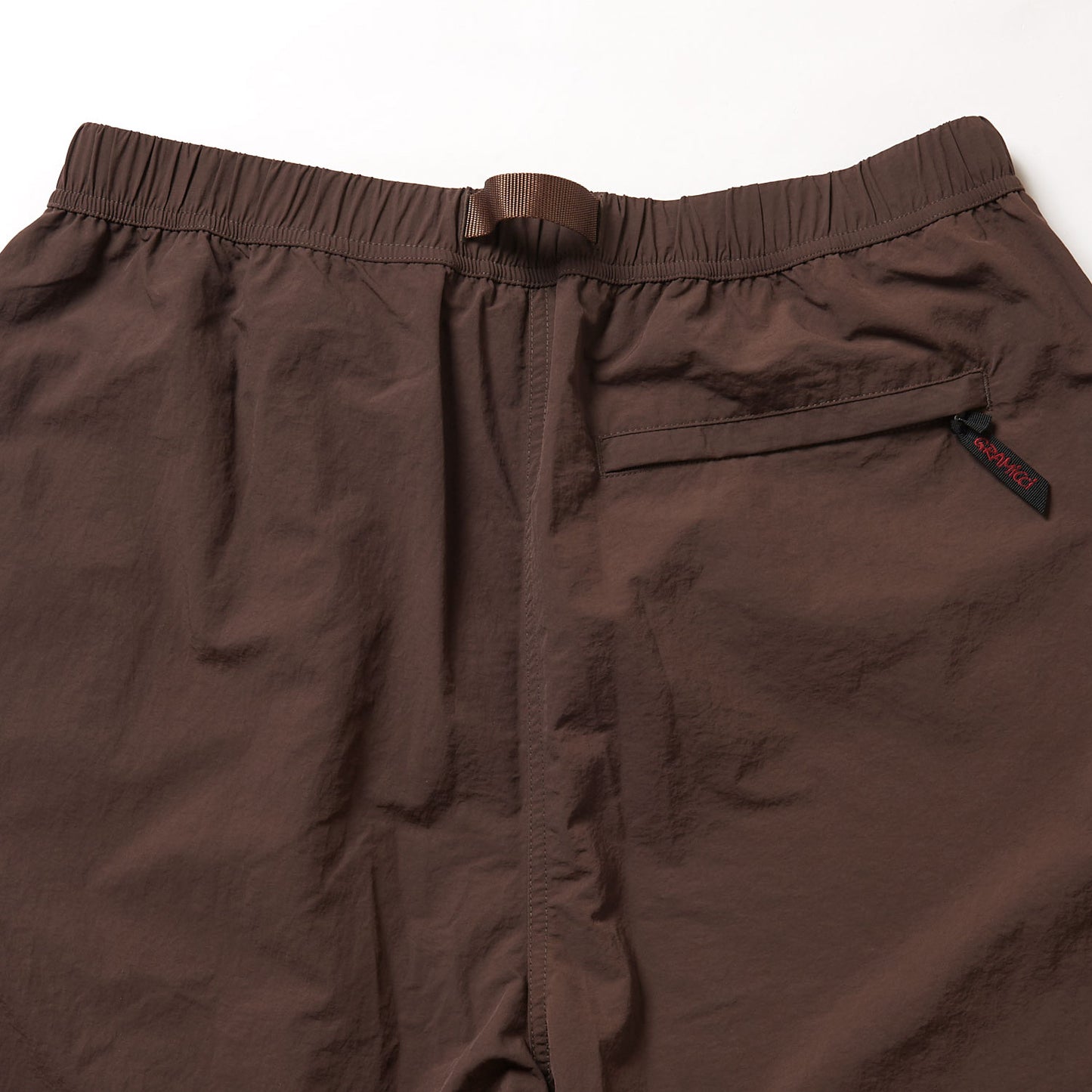 The Gramicci Nylon Runners are brown nylon pants with an elastic waistband, integrated belt, and a single back pocket accented by a small black and red woven tag.
