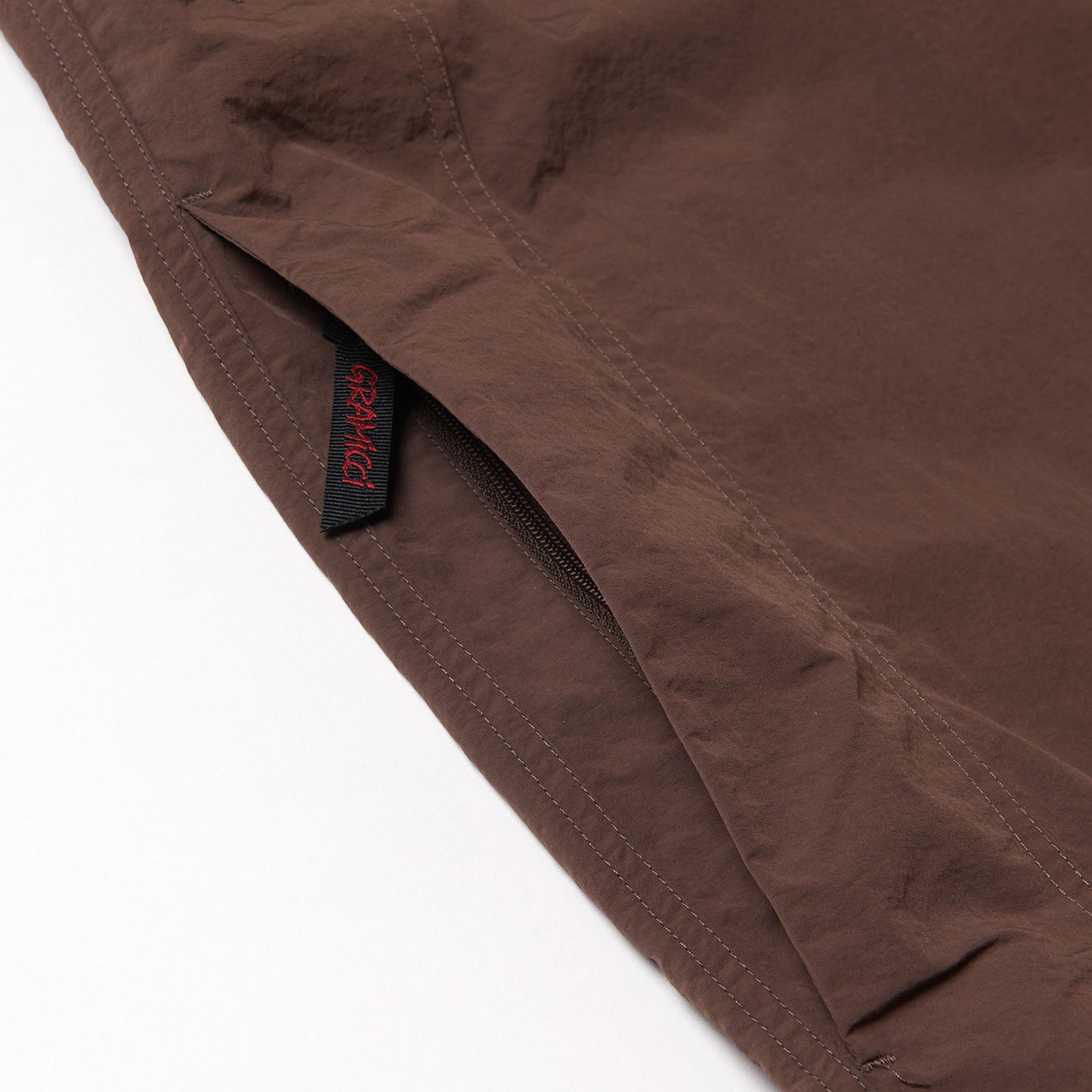 Close-up of Gramicci Nylon Runners in brown, highlighting a side seam zipper pocket with a black and red Gramicci tag stitched into the seam.
