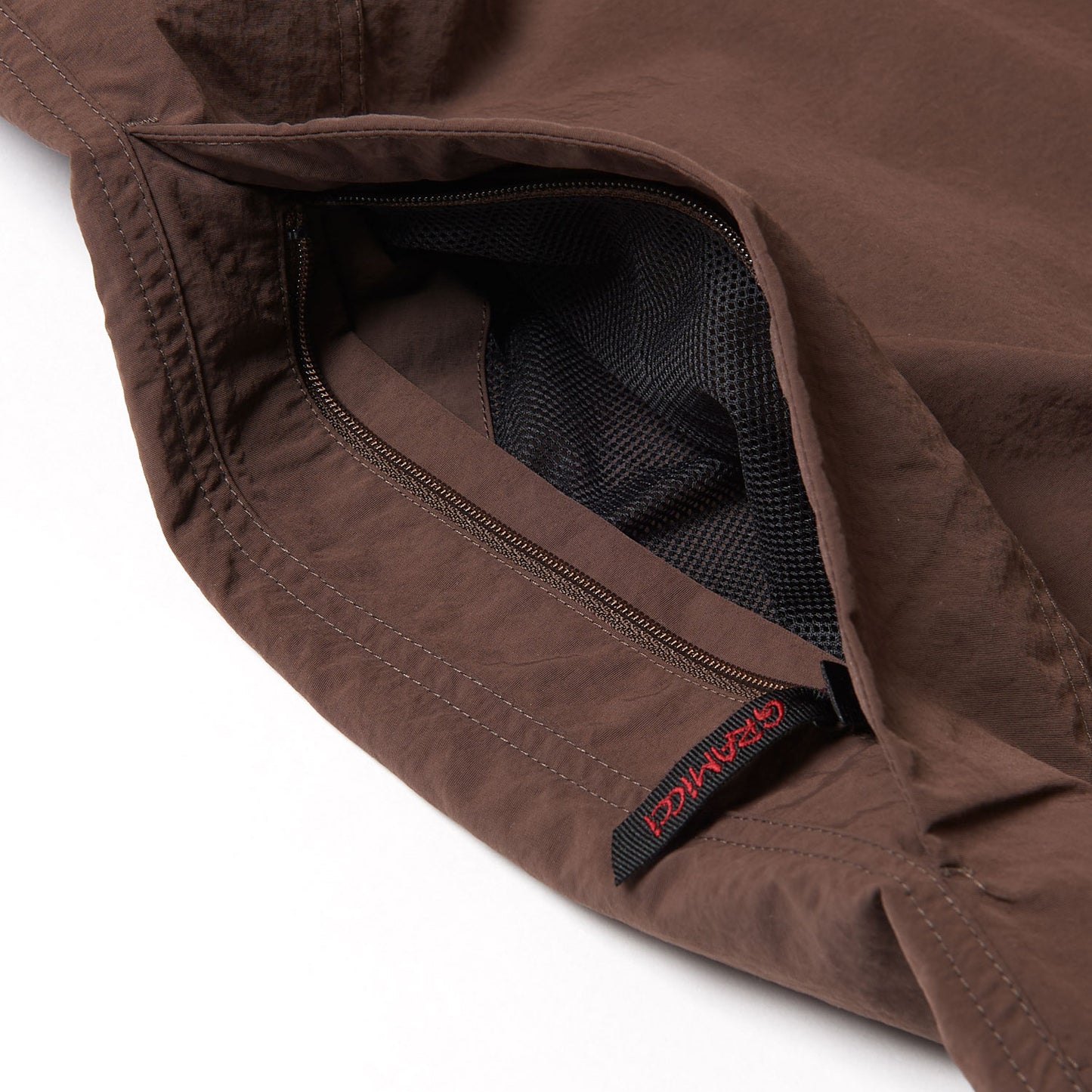 Close-up of Gramicci Nylon Runners showing a brown fabric with a zippered pocket, black mesh lining, and a black tag with red lettering.