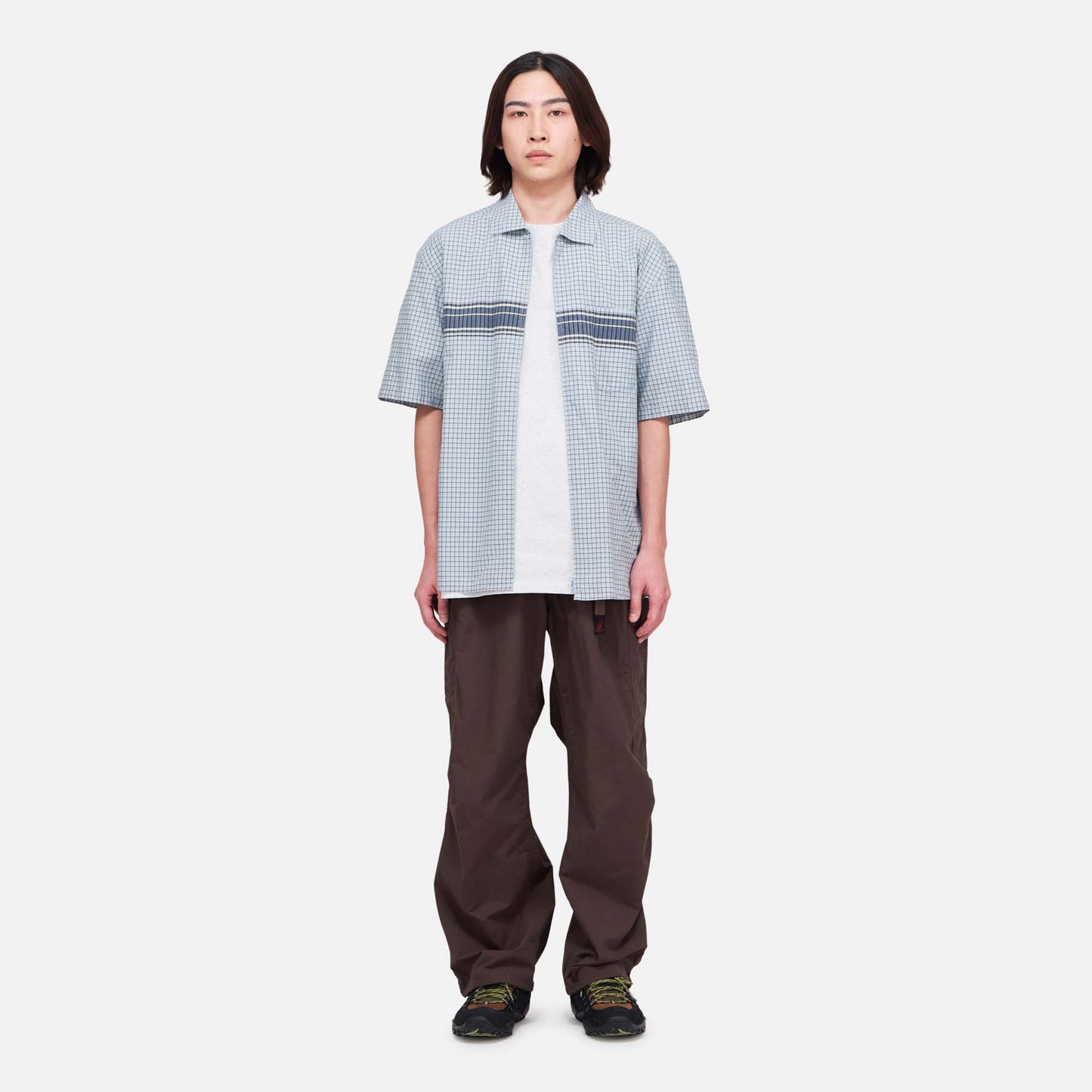 A person faces forward, wearing Gramicci Nylon Runners in brown, a light blue short-sleeve shirt over a white t-shirt, and black shoes, all set against a plain white background.