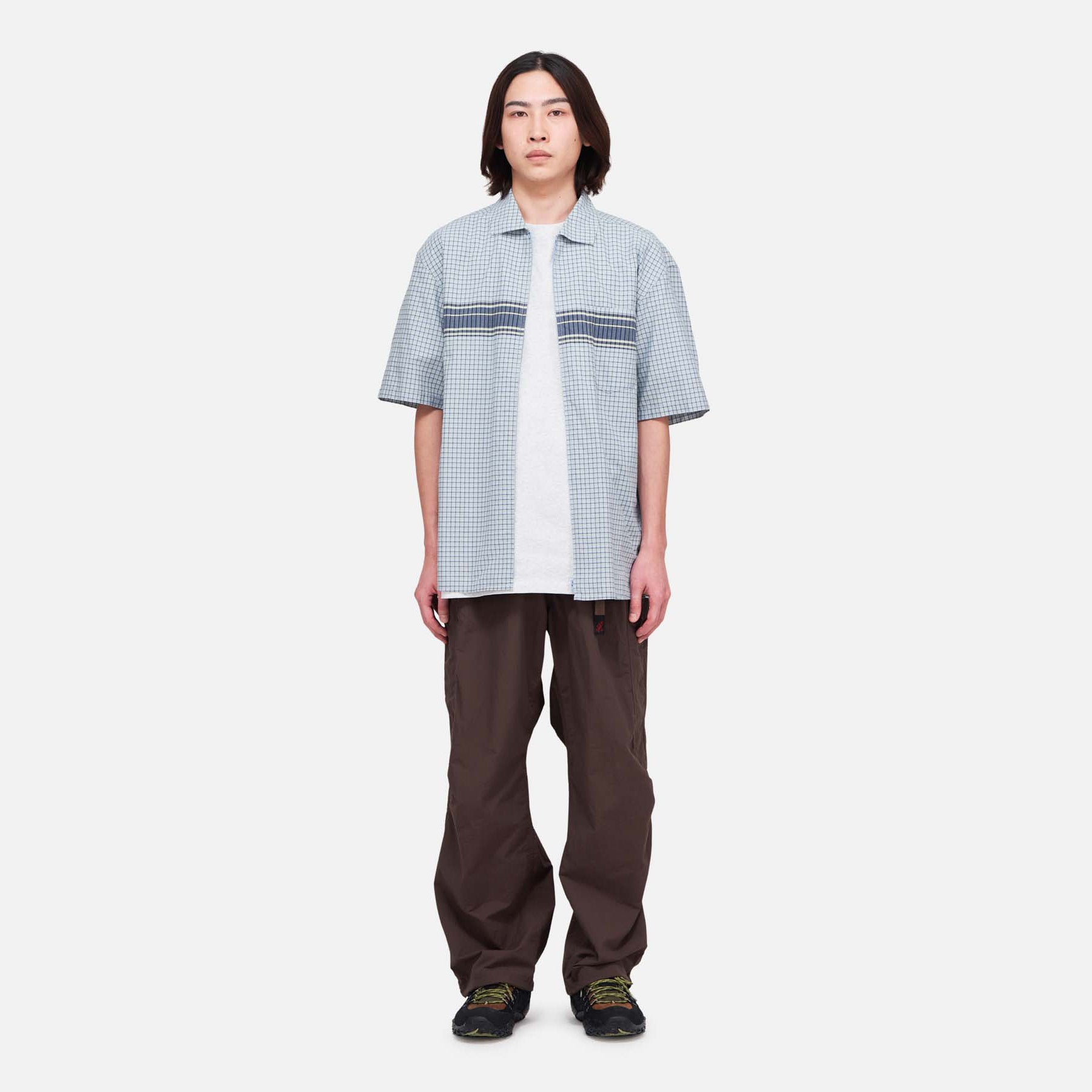 A person faces forward, wearing Gramicci Nylon Runners in brown, a light blue short-sleeve shirt over a white t-shirt, and black shoes, all set against a plain white background.