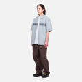 A person faces slightly left against a white background, wearing loose Gramicci Nylon Runners in brown, a short-sleeve plaid shirt over a white T-shirt, and black shoes.