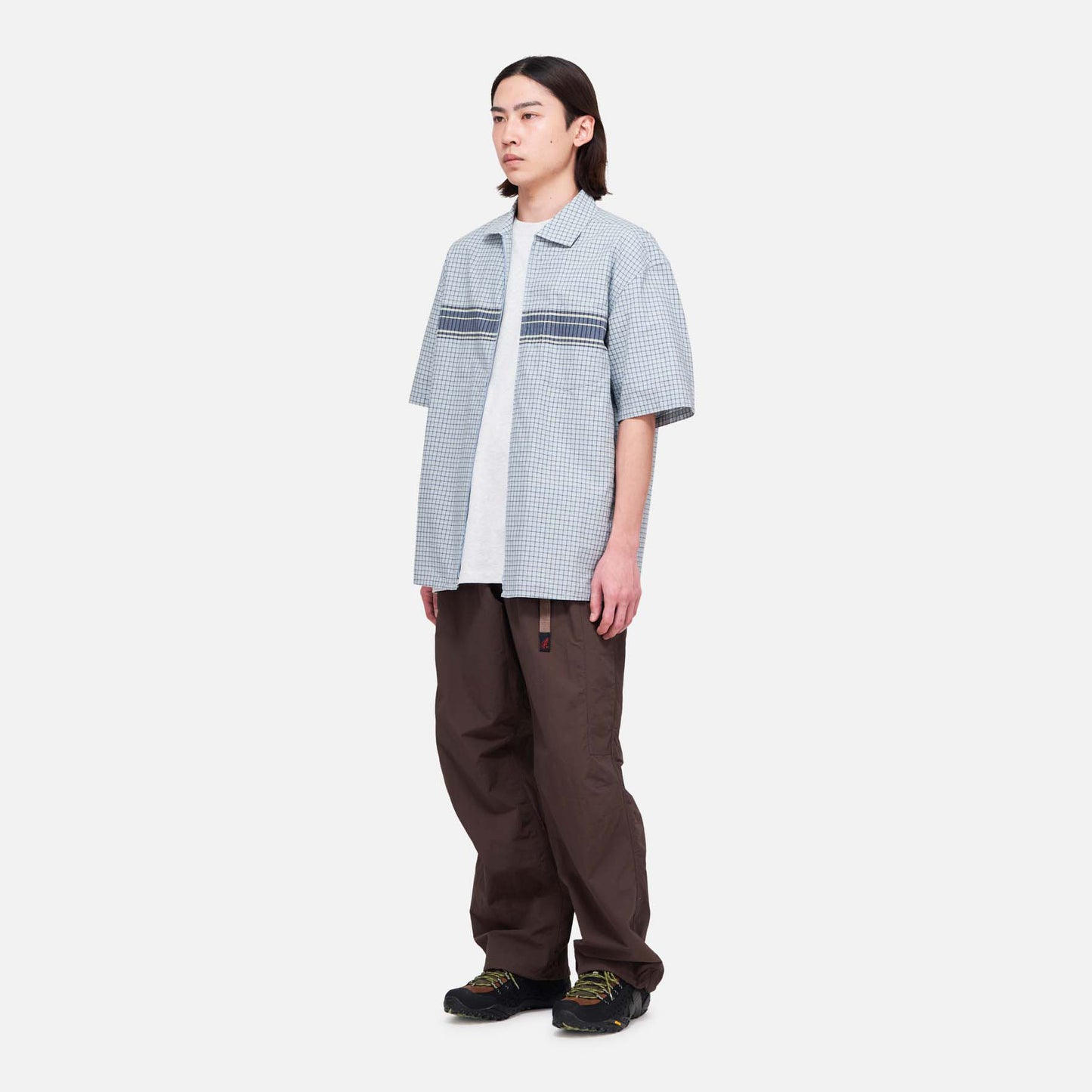 A person faces slightly left against a white background, wearing loose Gramicci Nylon Runners in brown, a short-sleeve plaid shirt over a white T-shirt, and black shoes.