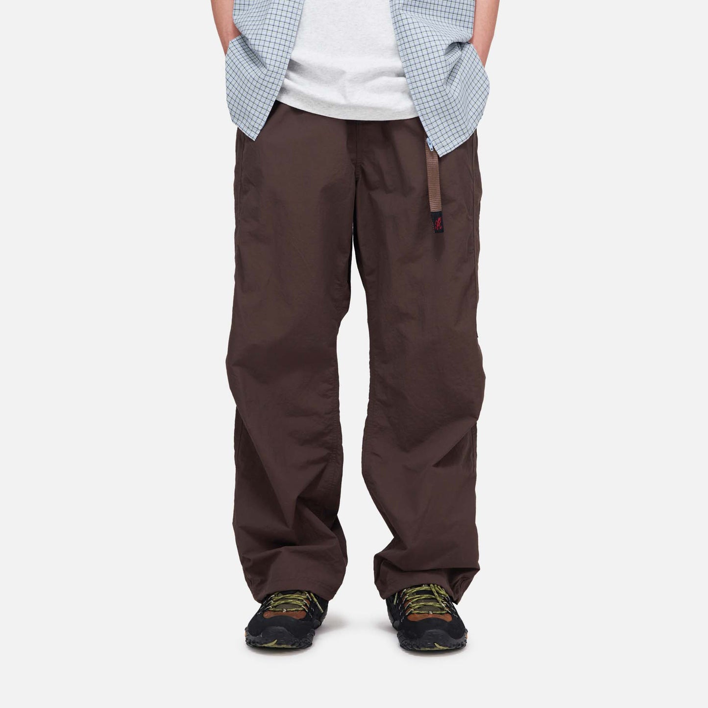 A person is wearing loose-fitting brown Gramicci Nylon Runners, black sneakers with green details, a white t-shirt, and a blue checkered shirt. The person's head is not visible.