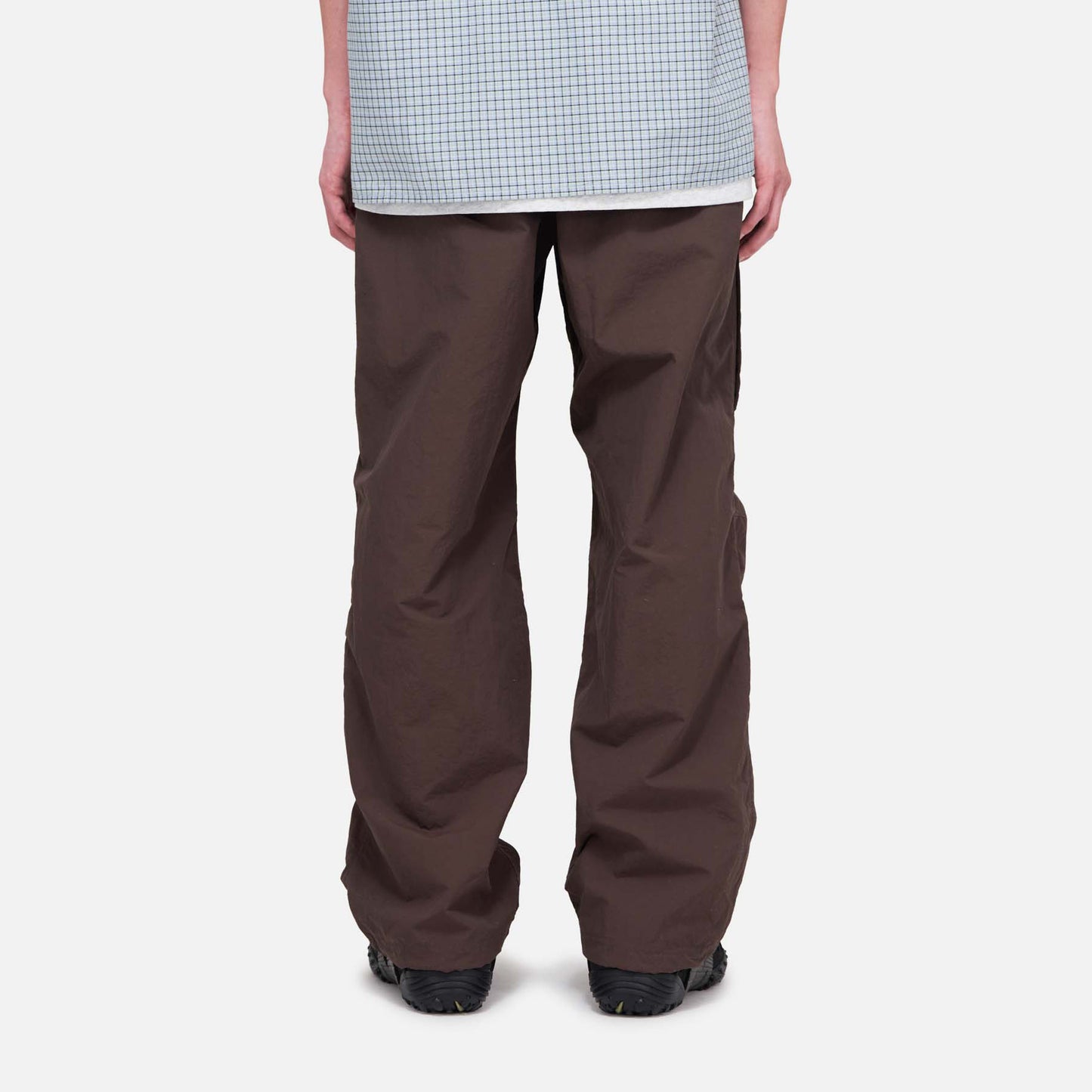 A person is seen from the back wearing Gramicci Nylon Runners, loose brown pants, and a light blue checkered shirt, standing against a plain white background.