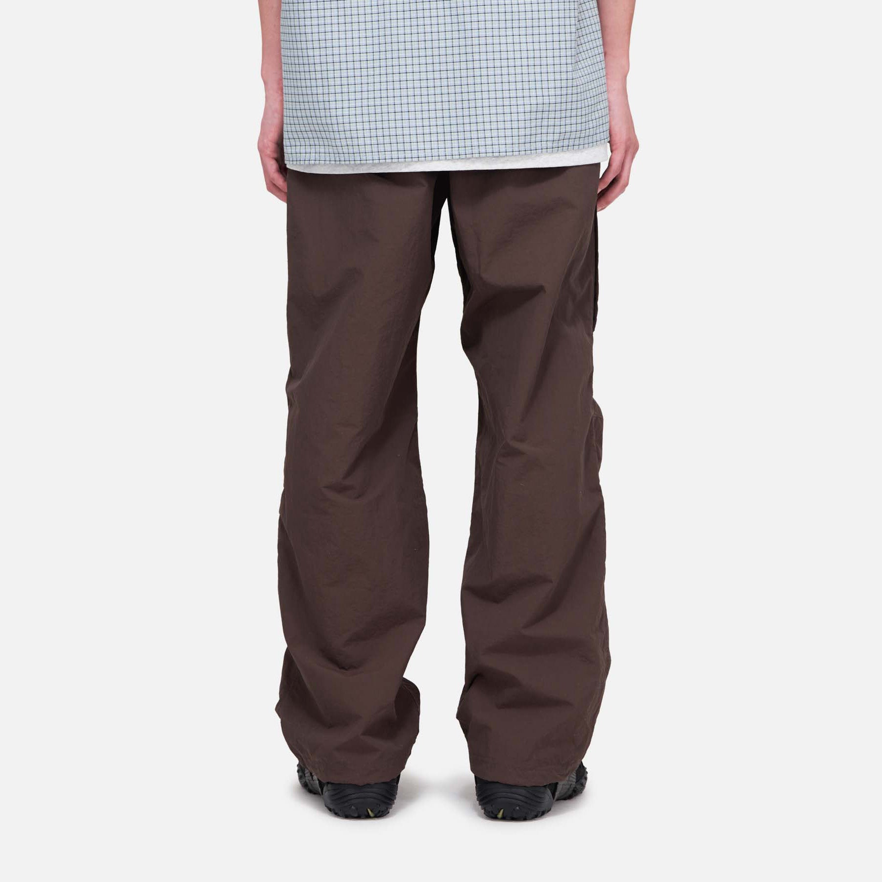 A person is seen from the back wearing Gramicci Nylon Runners, loose brown pants, and a light blue checkered shirt, standing against a plain white background.
