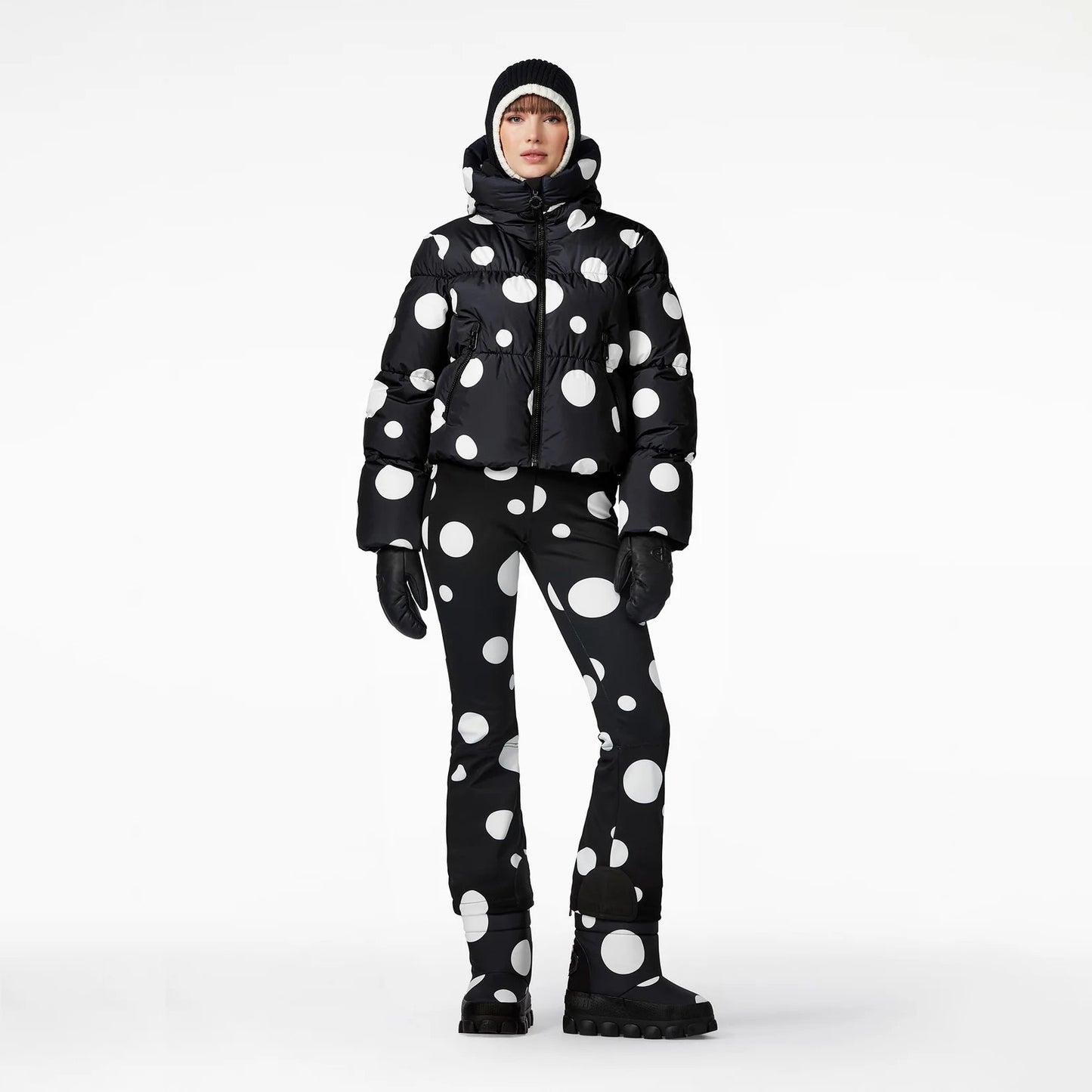 Dressed in Goldbergh's Grand Ski Jacket in Dot Black, with coordinating pants, beanie, and gloves, a person stands against a plain background.
