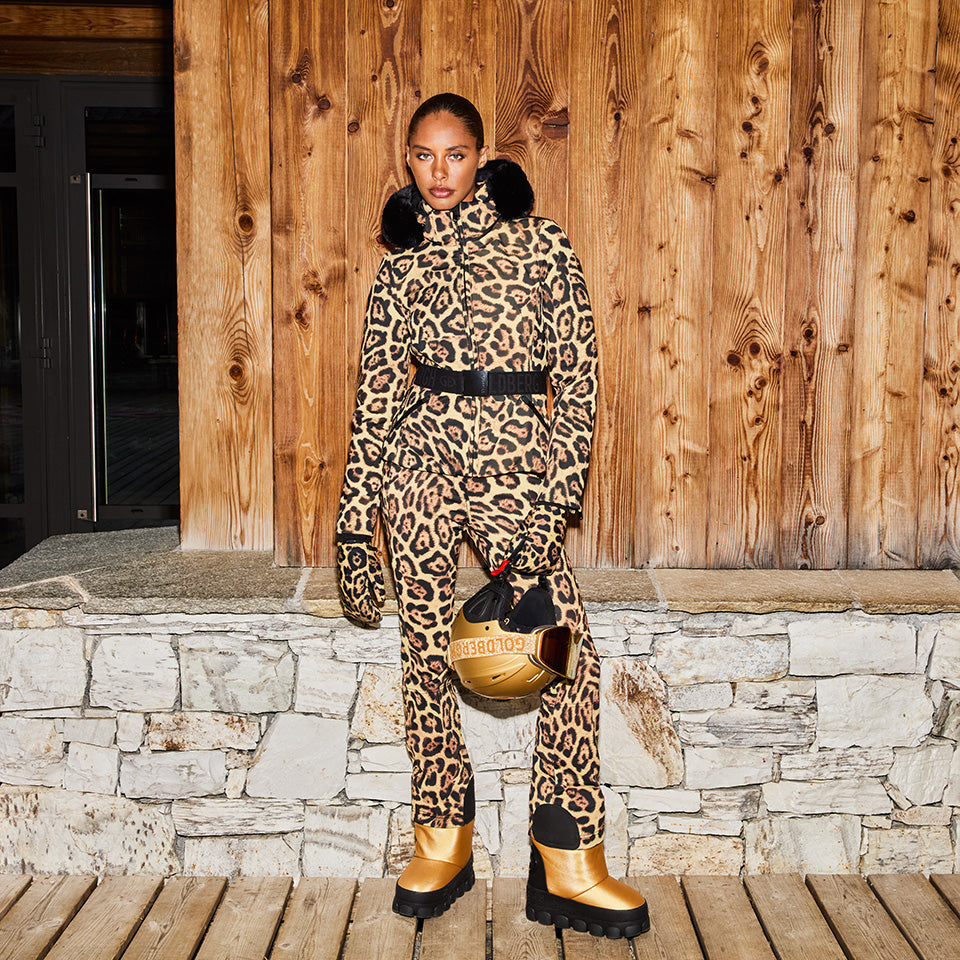 A person stands against a wooden wall wearing Goldbergh's breathable Minou Ski Pants in a leopard print (Jaguar), accessorized with a black belt, gloves, and gold boots while holding a matching gold helmet with black details.