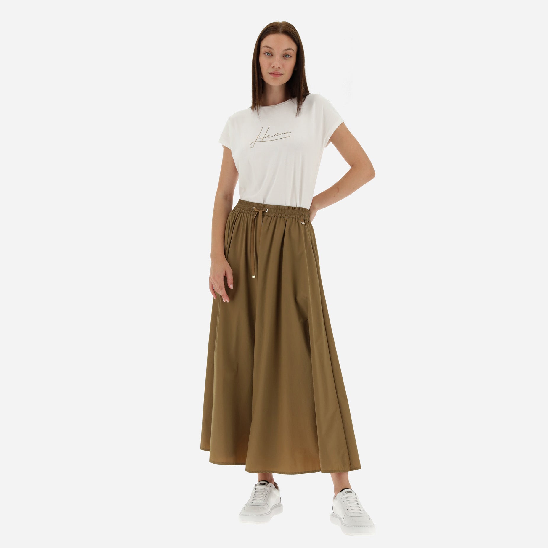 A woman stands against a plain background wearing a white t-shirt with cursive print, the Herno Nylon Stretch Skirt in Camel featuring an elastic waist insert, and white sneakers.