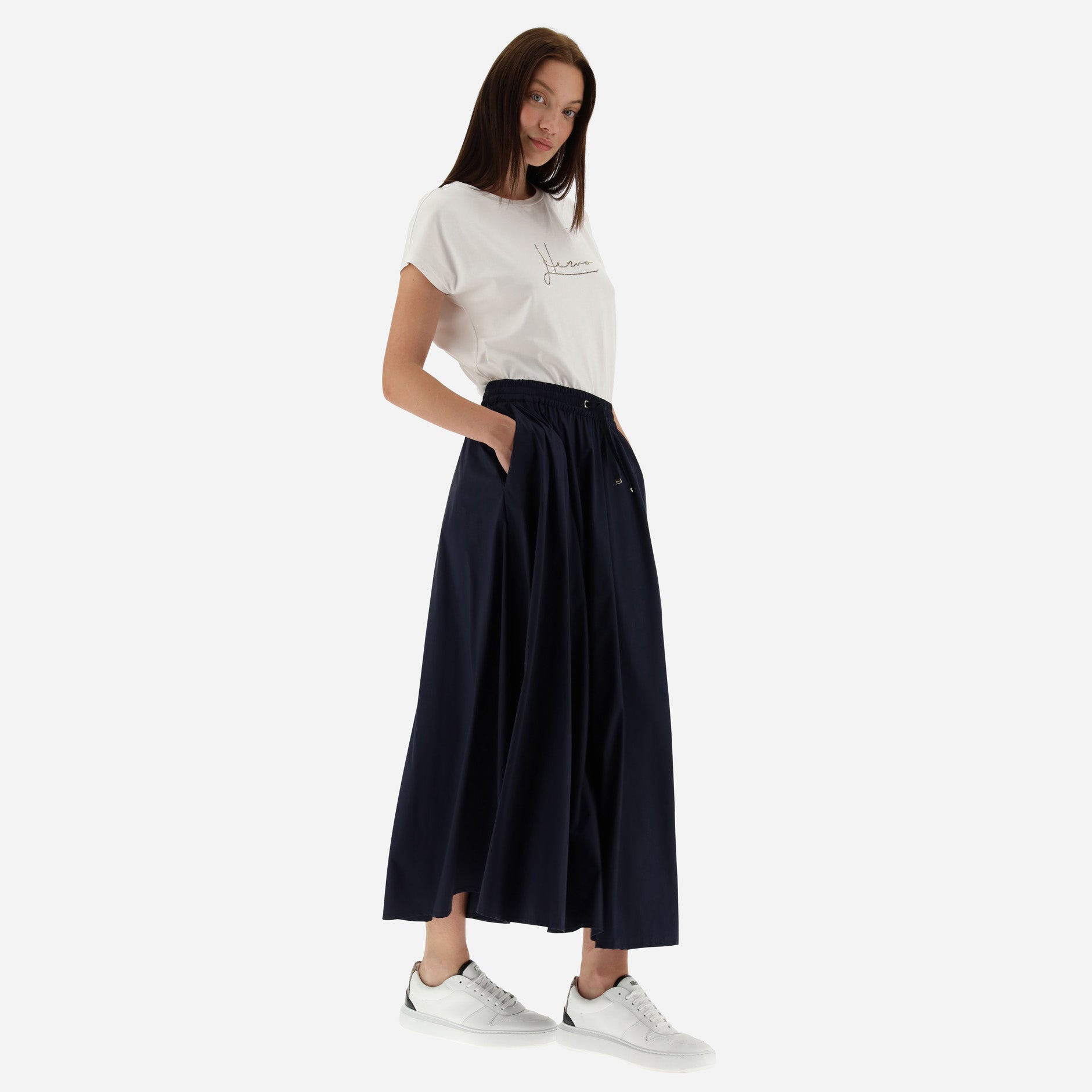 A woman stands with hands in pockets, wearing a white T-shirt, white sneakers, and the Herno Nylon Stretch Skirt in navy with an elastic waist against a plain background.