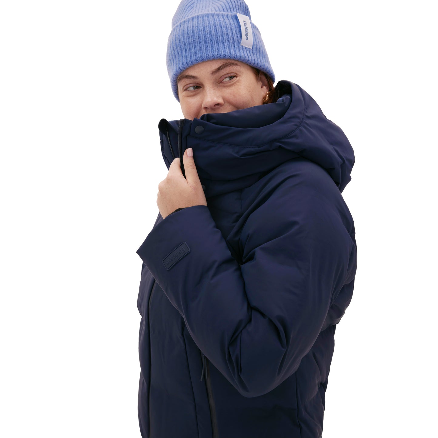 A person wearing a Georgie Puffer Jacket by Halfdays in navy with mountain performance features and a light blue beanie stands on a white background, looking to the side.
