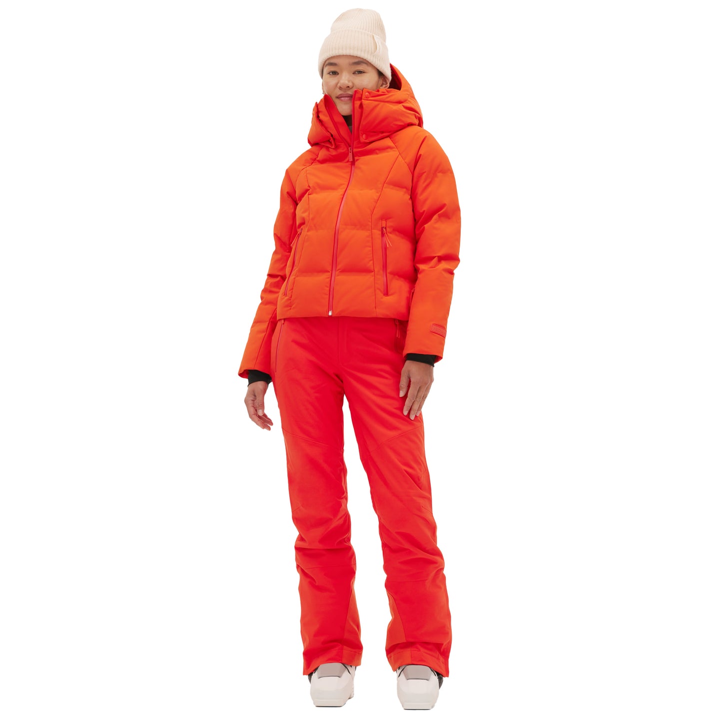A person in a Georgie Puffer Jacket by Halfdays and ski pants with a beige beanie and white boots stands against a plain white backdrop, ready for mountain performance.