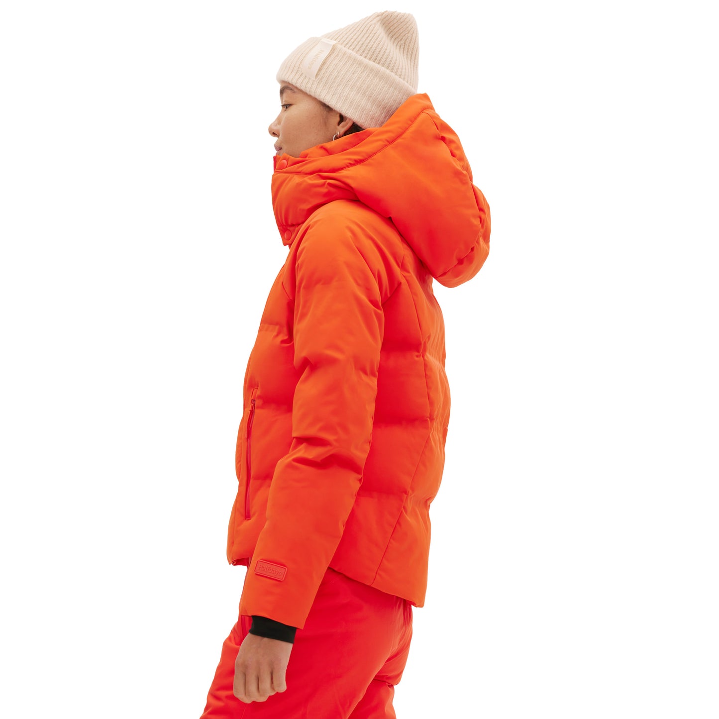 A person wearing the orange Georgie Puffer Jacket by Halfdays and a white knit hat stands sideways against a white background, highlighting their mountain performance readiness.