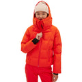 Dressed for the slopes, a person stands on a white backdrop wearing the Georgie Puffer Jacket by Halfdays, an orange ski jacket with mountain performance features and a hood, paired with a beige beanie.