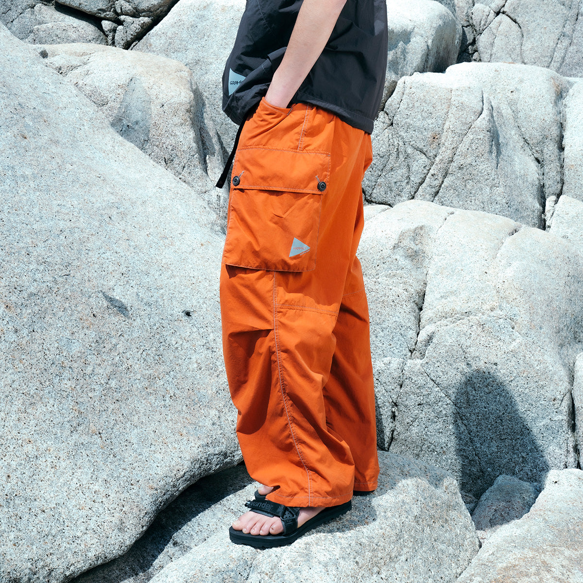 An individual wearing Gramicci x And Wander Military Wide Pants and sandals stands on large gray rocks, facing left.