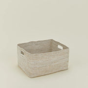 The Hawkins New York Essential Low Basket, a rectangular handwoven seagrass design with two handles, provides stylish blanket storage and reduces clutter on a simple light-colored surface.