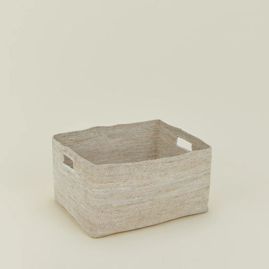 The Hawkins New York Essential Low Basket, a rectangular handwoven seagrass design with two handles, provides stylish blanket storage and reduces clutter on a simple light-colored surface.