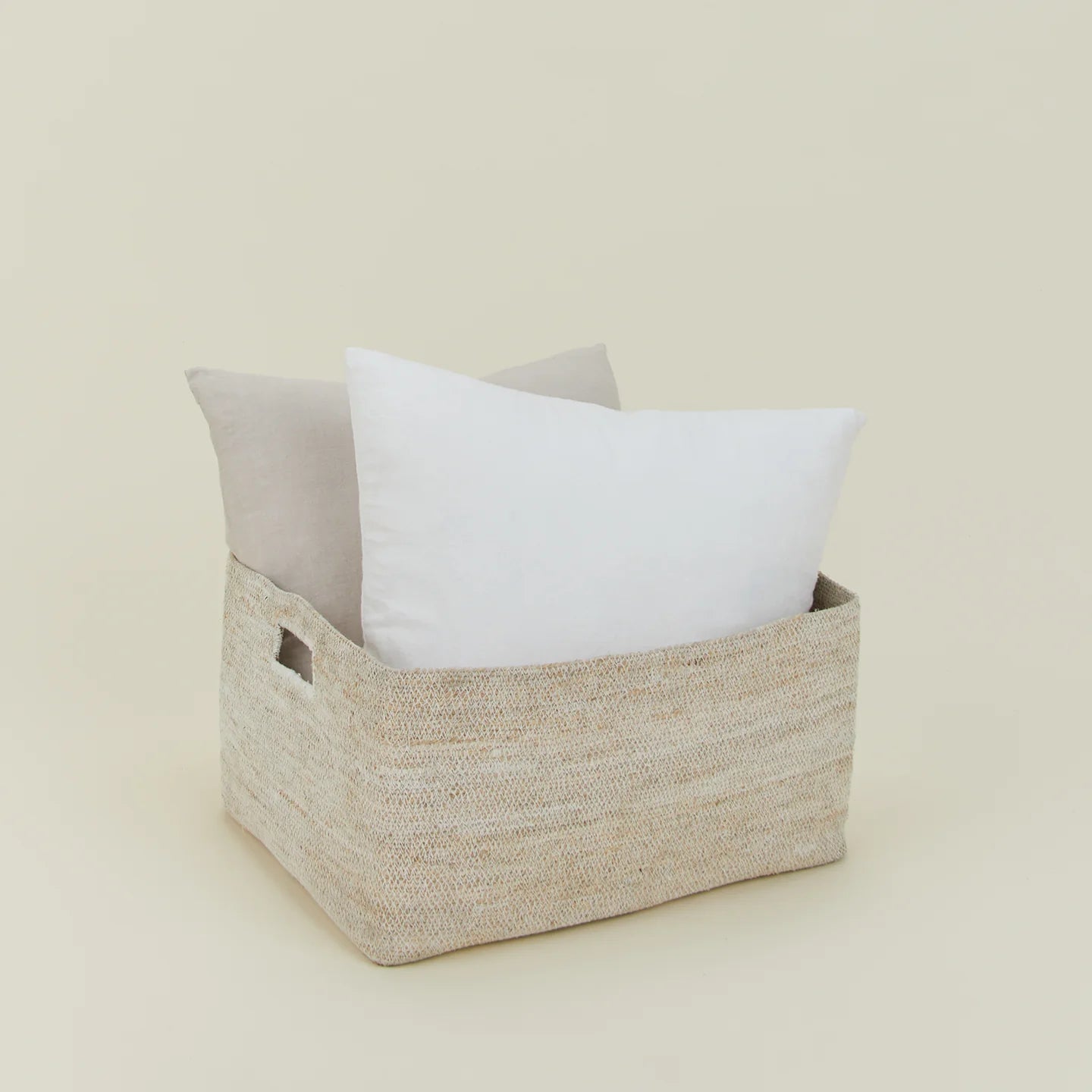 The Essential Low Basket by Hawkins New York, handwoven from seagrass, neatly holds a white pillow and a beige pillow against a simple light background, making it ideal for decluttering any space.
