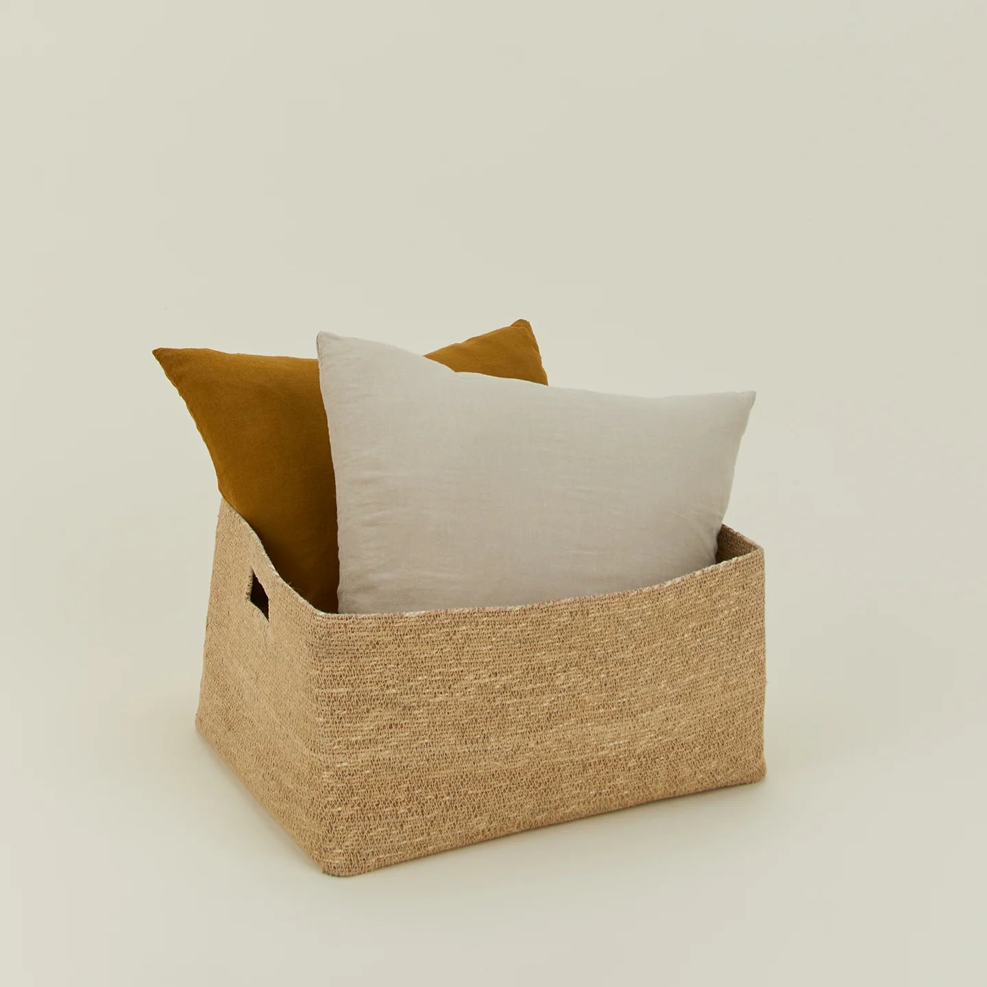 The Essential Low Basket by Hawkins New York, handwoven from seagrass, comes with two large throw pillows—one white and one brown—for stylish blanket storage and clutter reduction against a plain light background.