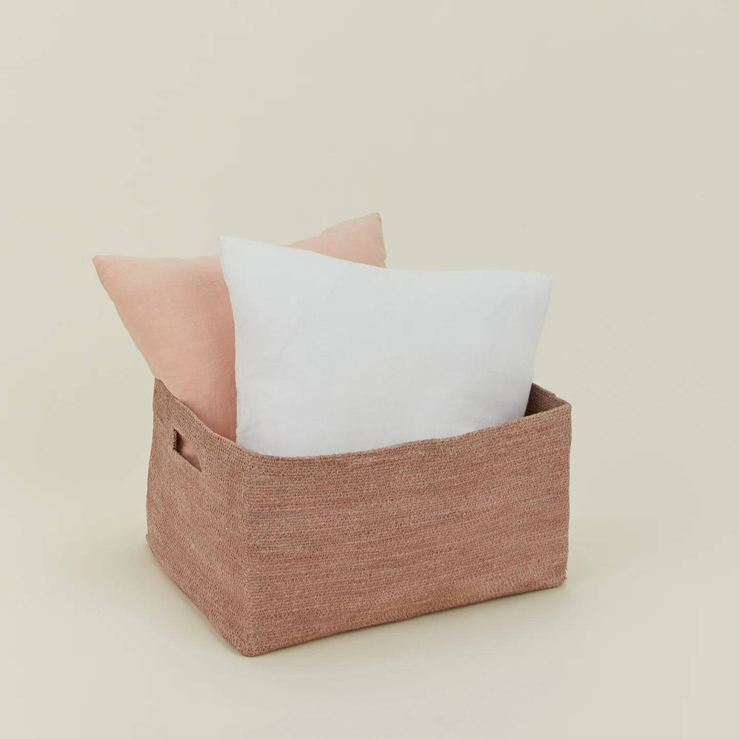 The Essential Low Basket from Hawkins New York, handwoven from seagrass, holds a white and light pink pillow—ideal for storing blankets or reducing clutter—set against a plain beige backdrop.