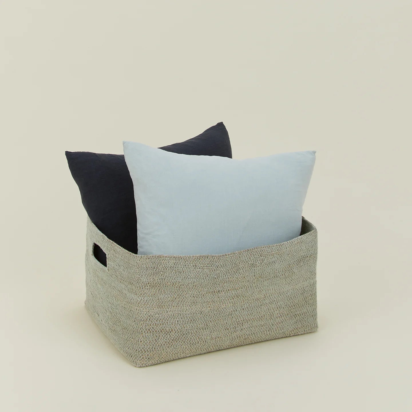 The Essential Low Basket by Hawkins New York, handwoven from seagrass and holding a light and dark blue pillow, is set against a plain light background—perfect for minimizing clutter.