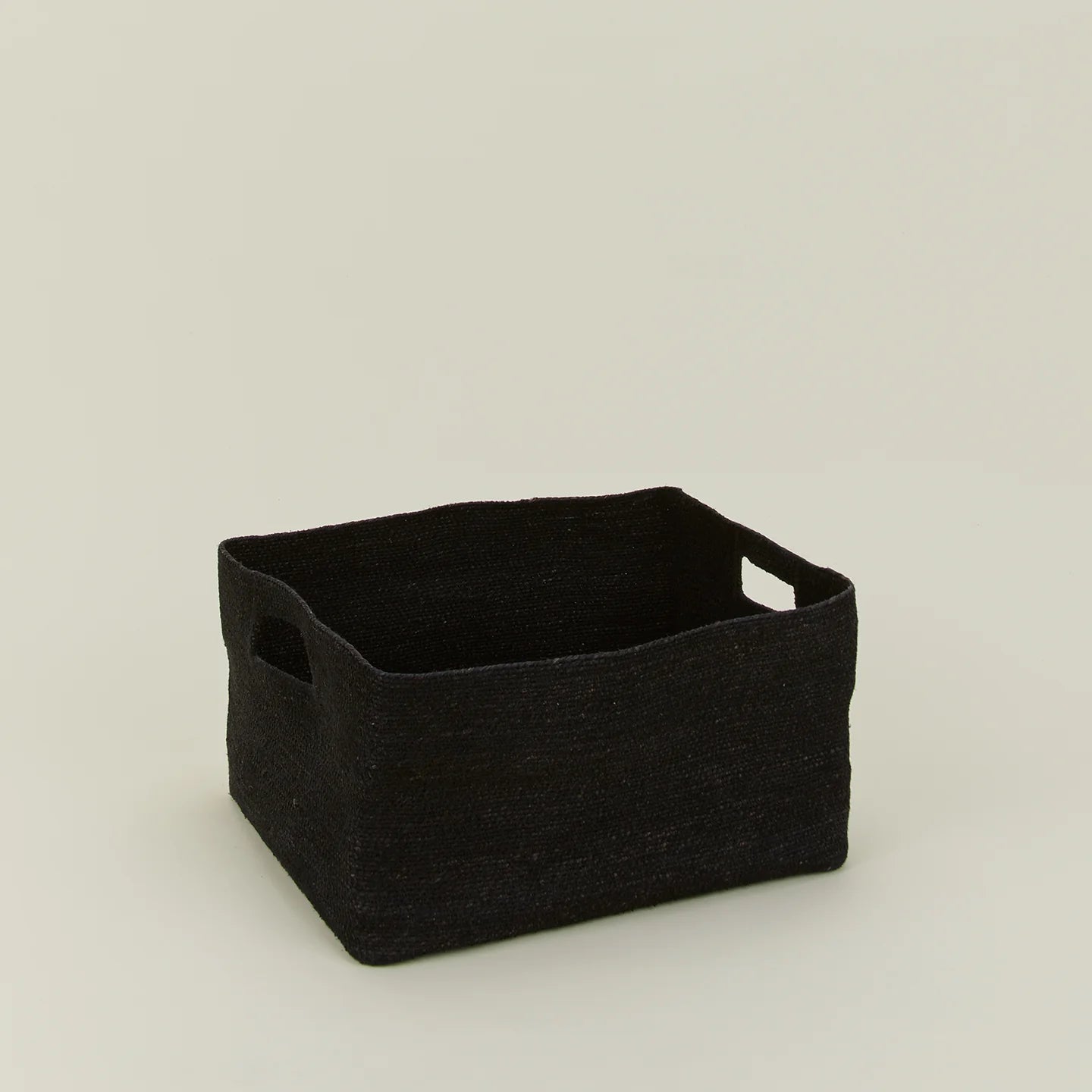 The Essential Low Basket by Hawkins New York is a handwoven black rectangular seagrass basket with side handles, ideal for storing blankets and reducing clutter, shown on a plain light-colored surface.