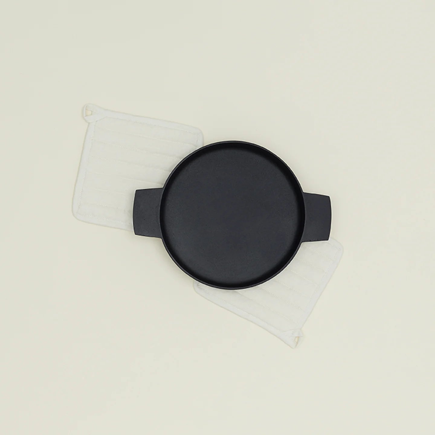 A round black baking dish with handles sits on two ivory Simple Linen Pot Holders by Hawkins New York, offering extra padding against a plain light background.
