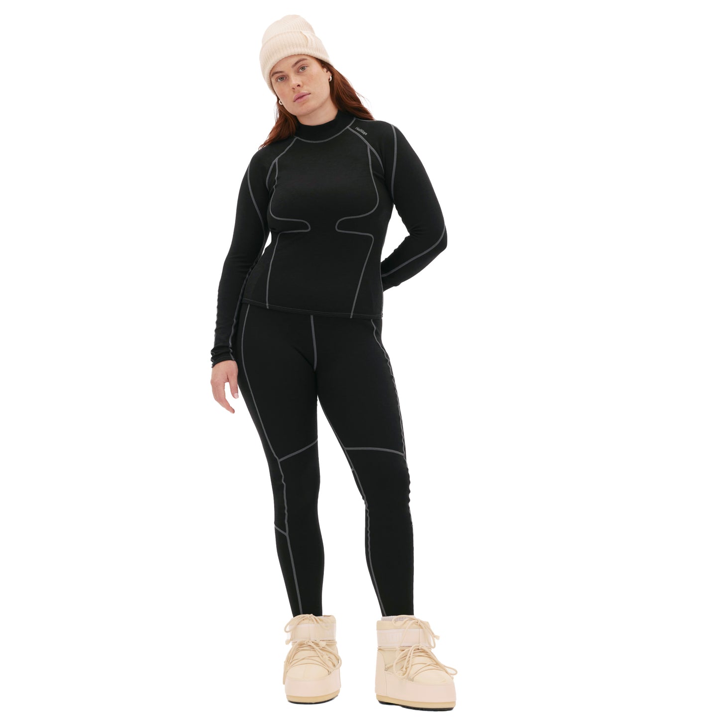 A person in Hallie Merino Legging by Halfdays, a black thermal midweight blend outfit, pairs it with a beige knit hat and winter boots against a white background.