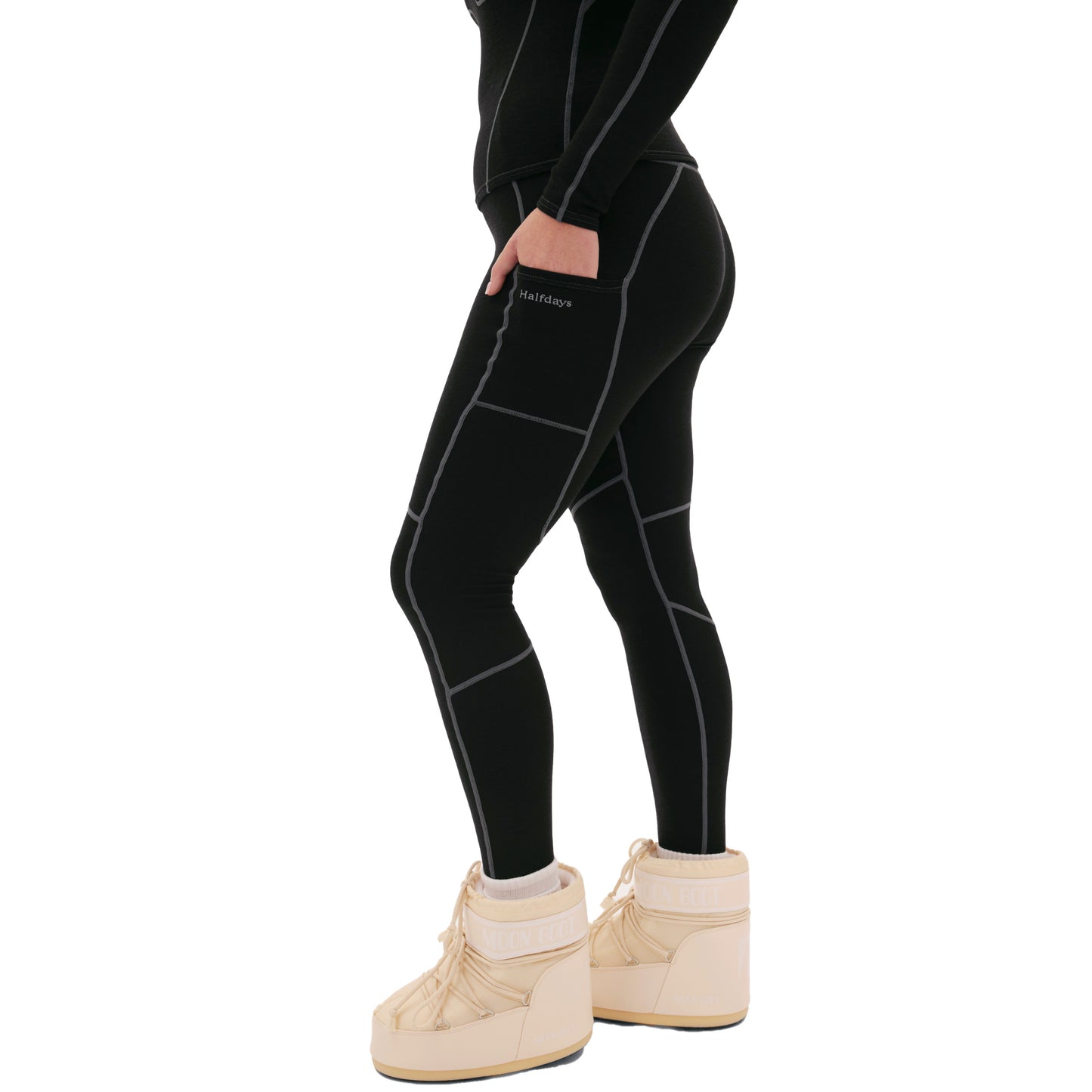The image shows a person from mid-thigh to feet wearing Halfdays' Hallie Merino Legging in black with white stitching, paired with beige snow boots. These cozy midweight leggings provide style and warmth for chilly days.