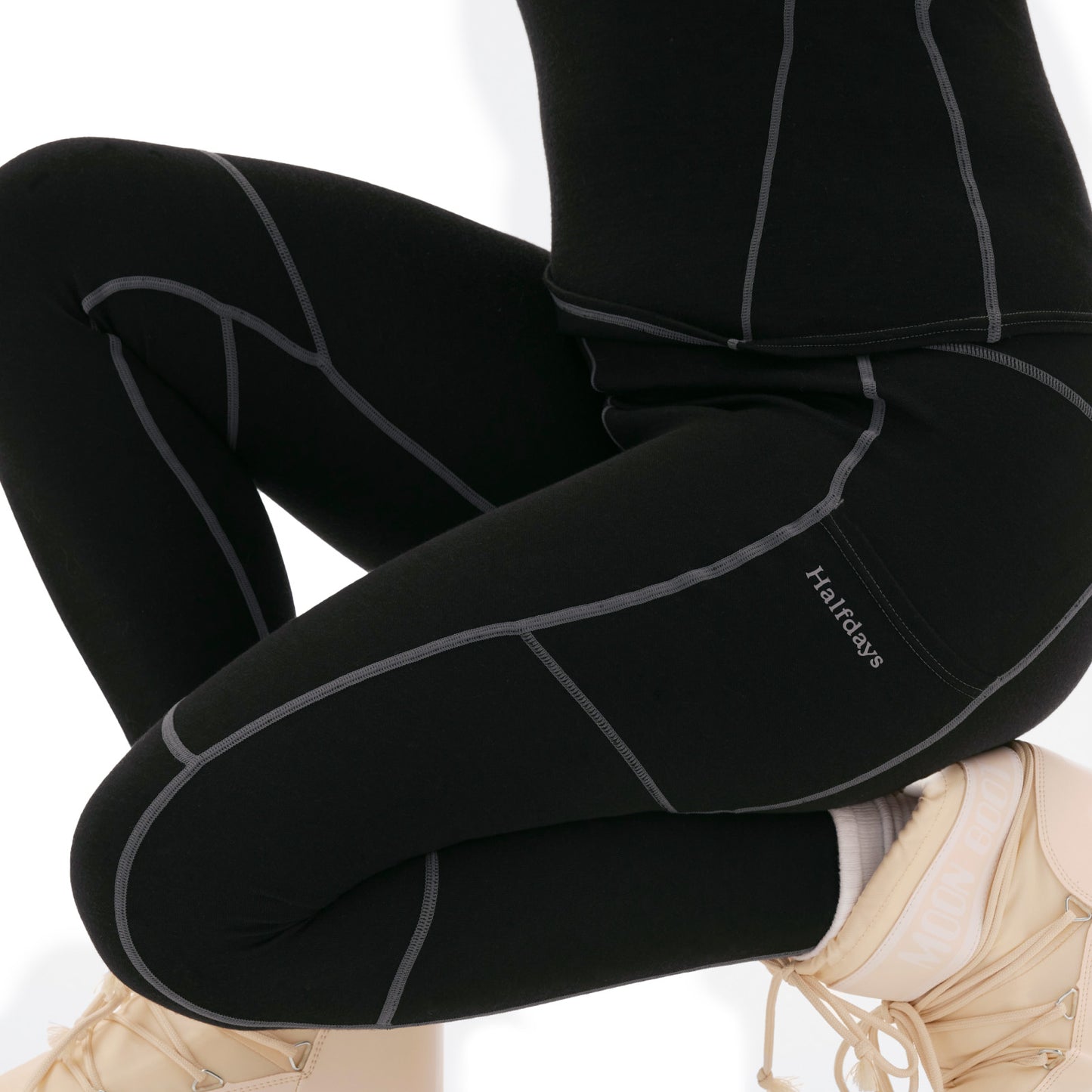 A person wearing black Halfdays leggings with gray seams, featuring the "Halifax" logo, paired with beige sneakers and a cozy Hallie merino wool base layer.
