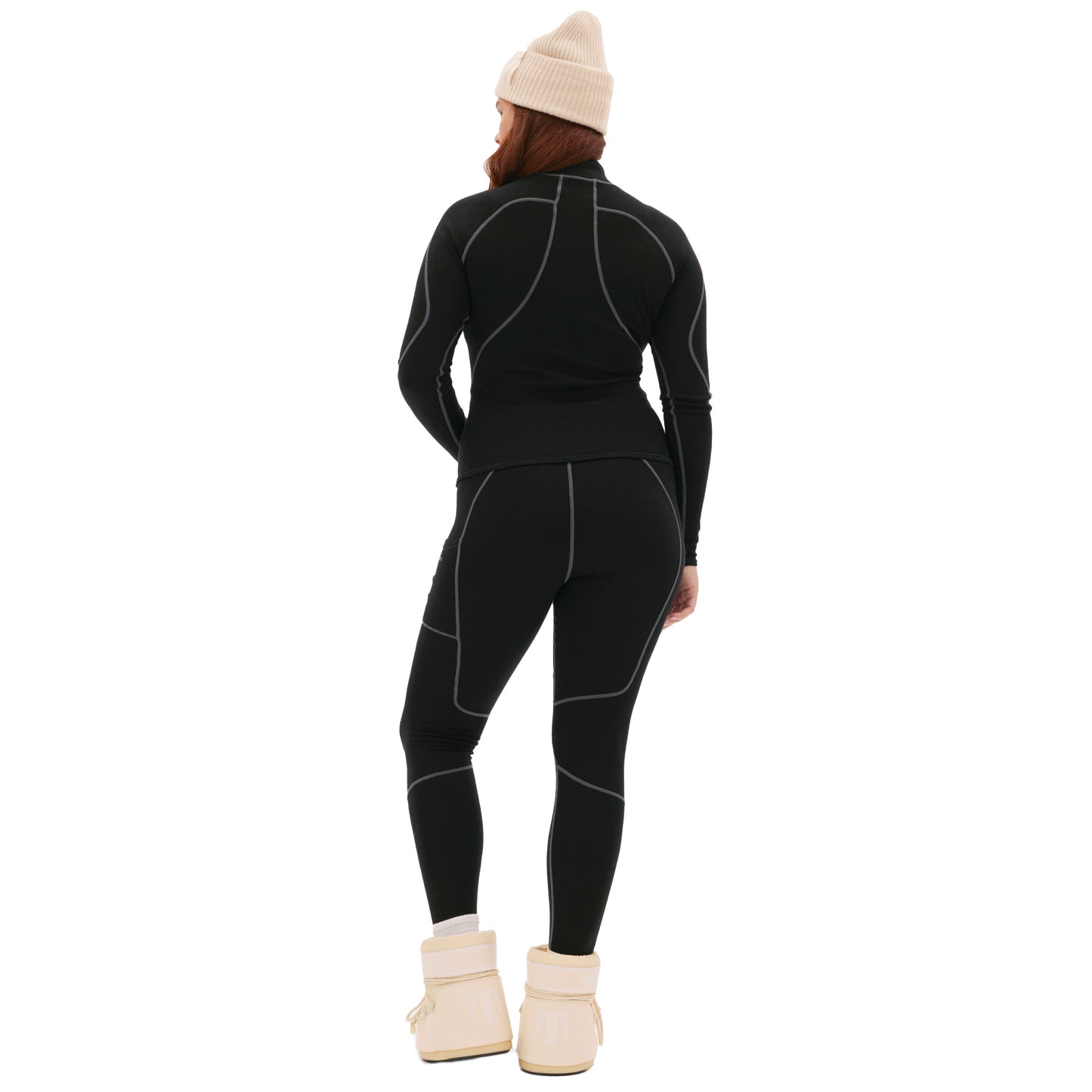 A person is seen from behind in a black athletic outfit with beige beanie and boots, against a plain backdrop. Hidden beneath their attire is the Hallie Merino Legging from Halfdays, providing cozy warmth and comfort.