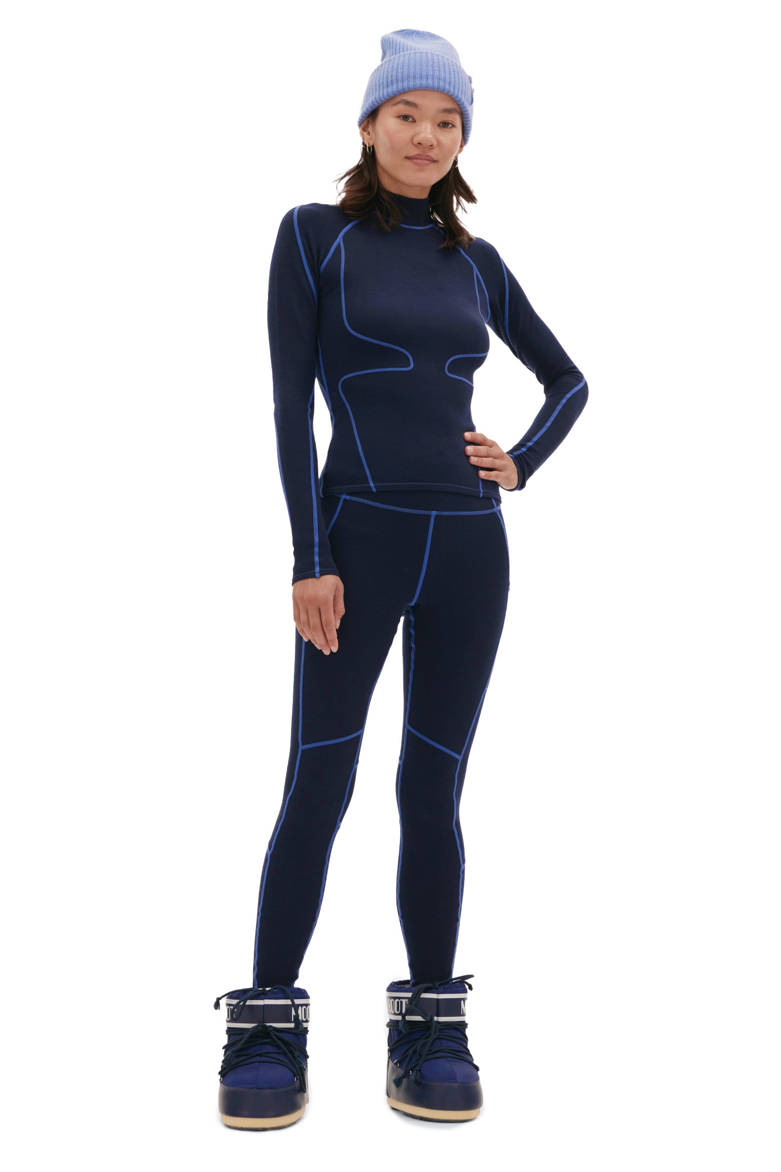 A person in a dark blue outfit with light blue accents and large snow boots stands looking forward. A light blue beanie completes the look, while Halfdays' Hallie Merino Legging peeks out, offering both warmth and style.