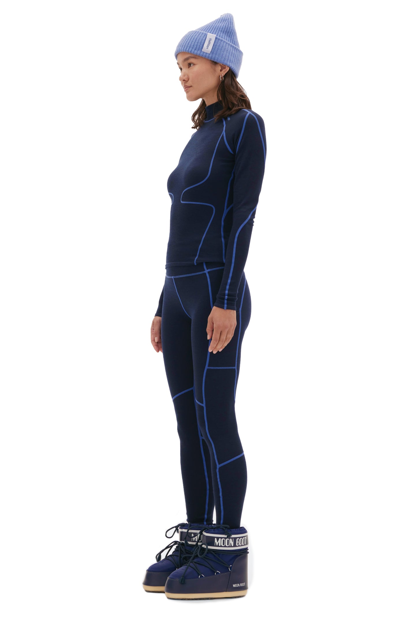 A person stands facing left, wearing a blue beanie, a dark blue midweight blend long-sleeve top, matching Halfdays Hallie Merino Legging with blue lines, and black and white boots.