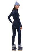 Against a white backdrop, the person wears a form-fitting navy outfit with light blue seams, paired with navy snow boots. A blue beanie completes the look, complemented by a warm Hallie Merino Legging base layer from Halfdays.