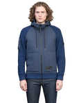A person with curly hair, wearing the luxurious blue Hedge Men's Performance Hoodie by Nobis, and jeans stands against a plain white background.