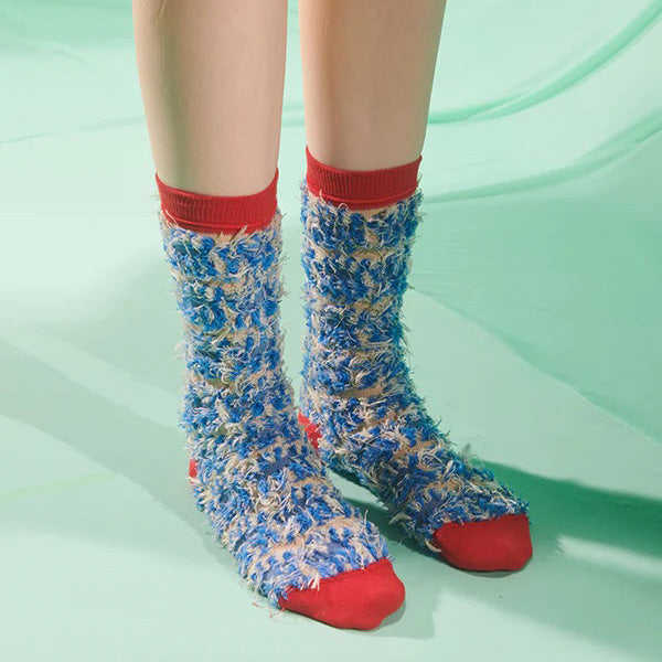 Someone stands on a light green surface wearing Henrik Vibskov Hairy Handsome Chair Socks Femme—blue and beige fuzzy socks with red cuffs, heels, toes, and an abstract pattern.