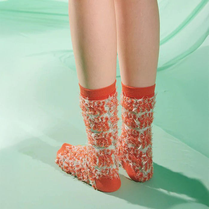 A person stands on a light green surface wearing Henrik Vibskov's Hairy Handsome Chair Socks Femme—orange and white socks with an abstract, fuzzy texture, echoing the brand’s playful aesthetic.