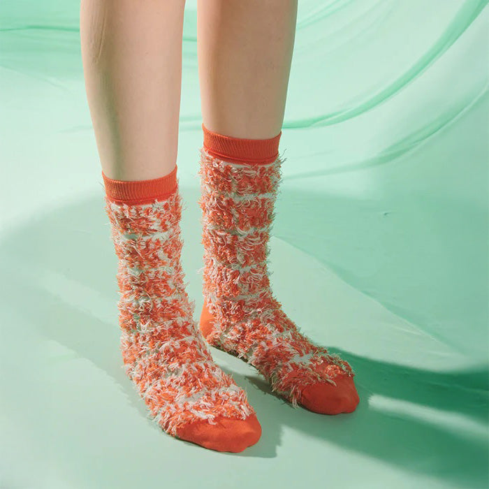A pair of feet in Henrik Vibskov’s Hairy Handsome Chair Socks Femme, featuring orange and white fuzzy abstract patterns, stand on a light green surface against a soft green background.