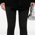 Against a plain backdrop, a person wears black textured leggings with an elastic waistband and a matching jacket while holding a silver bag. These chic and understated pieces might just be the sought-after Henrik Vibskov Honey Leggings.