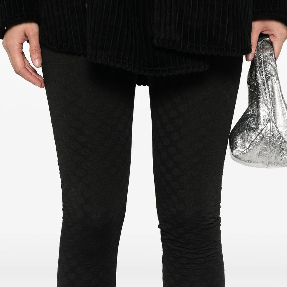 Against a plain backdrop, a person wears black textured leggings with an elastic waistband and a matching jacket while holding a silver bag. These chic and understated pieces might just be the sought-after Henrik Vibskov Honey Leggings.