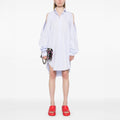 Wearing the Henrik Vibskov New Bumble Shirtdress in light blue with cold shoulders and long sleeves, a person stands against a white background, accessorized with a small black handbag and red slide sandals.