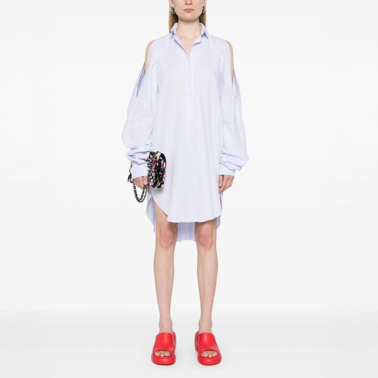 Wearing the Henrik Vibskov New Bumble Shirtdress in light blue with cold shoulders and long sleeves, a person stands against a white background, accessorized with a small black handbag and red slide sandals.