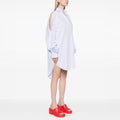 A woman in profile models the Henrik Vibskov New Bumble Shirtdress with open-shoulder cutouts, paired with bright red slide sandals, set against a plain white background.