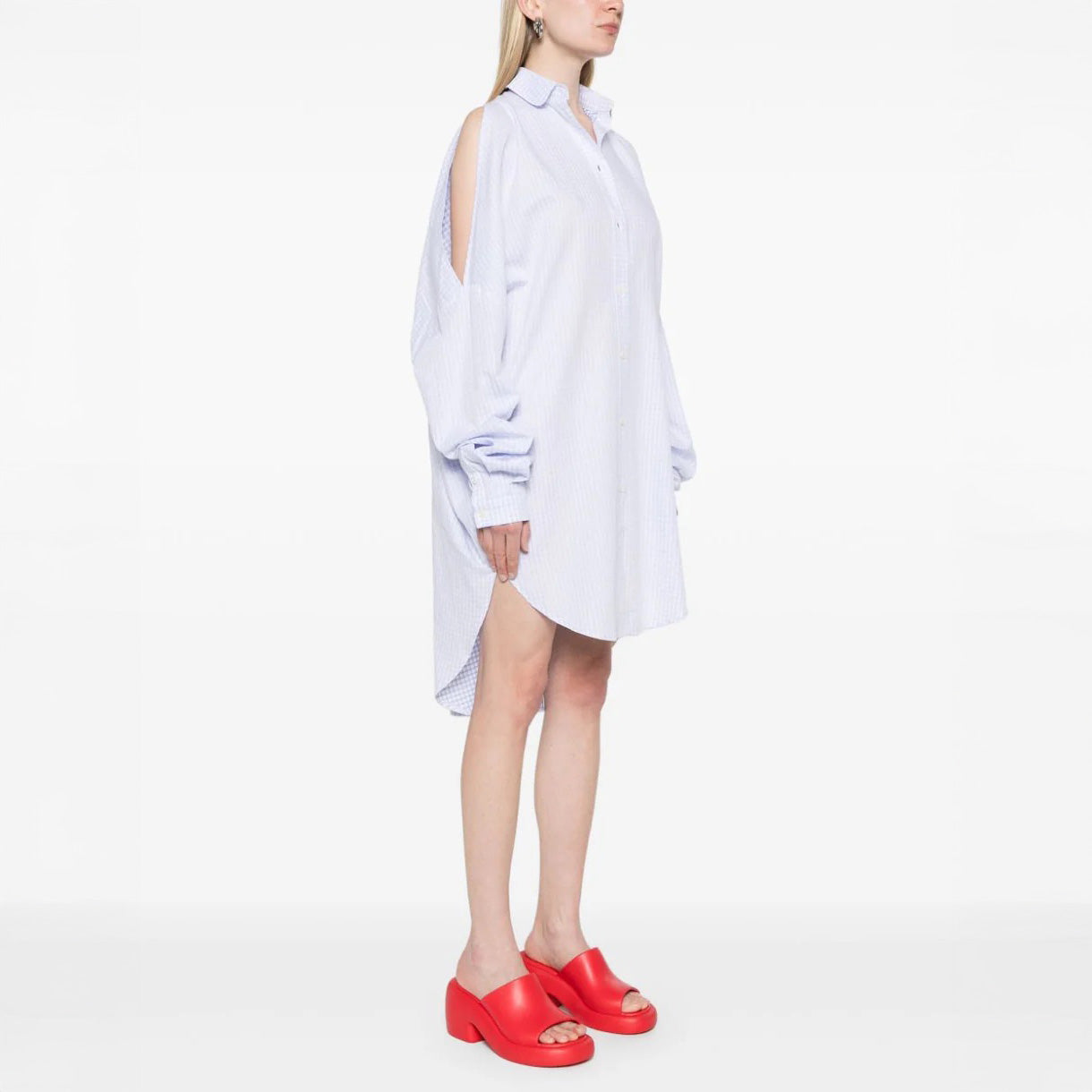 A woman in profile models the Henrik Vibskov New Bumble Shirtdress with open-shoulder cutouts, paired with bright red slide sandals, set against a plain white background.
