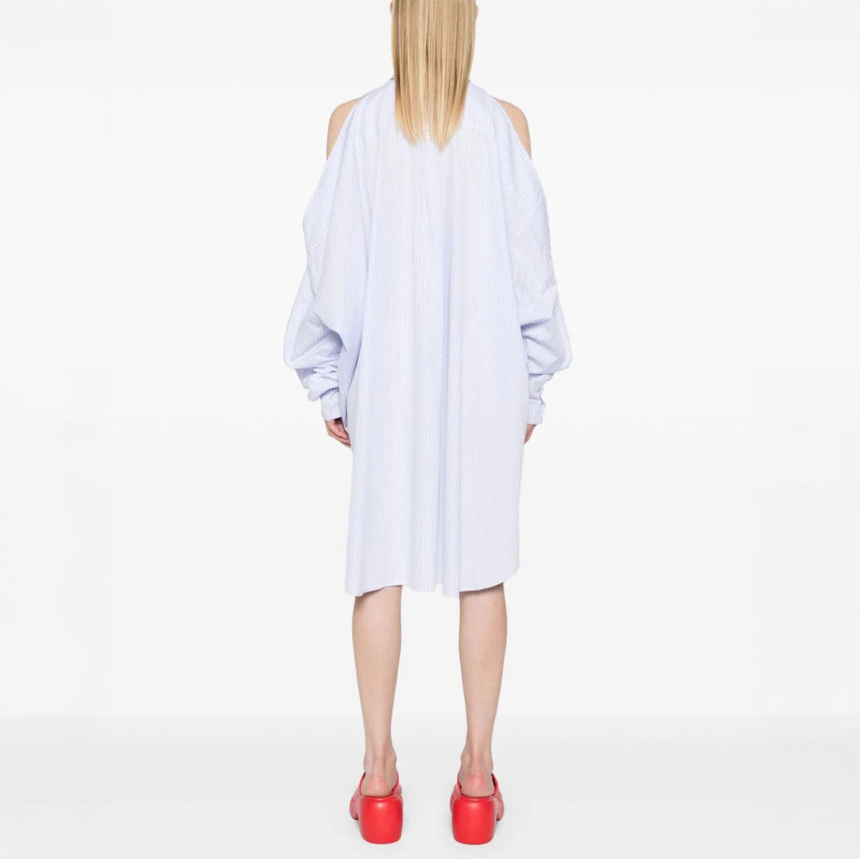 A person stands with back to camera in a loose light blue New Bumble Shirtdress by Henrik Vibskov, featuring shoulder cutouts, paired with bright red platform shoes against a plain white background.