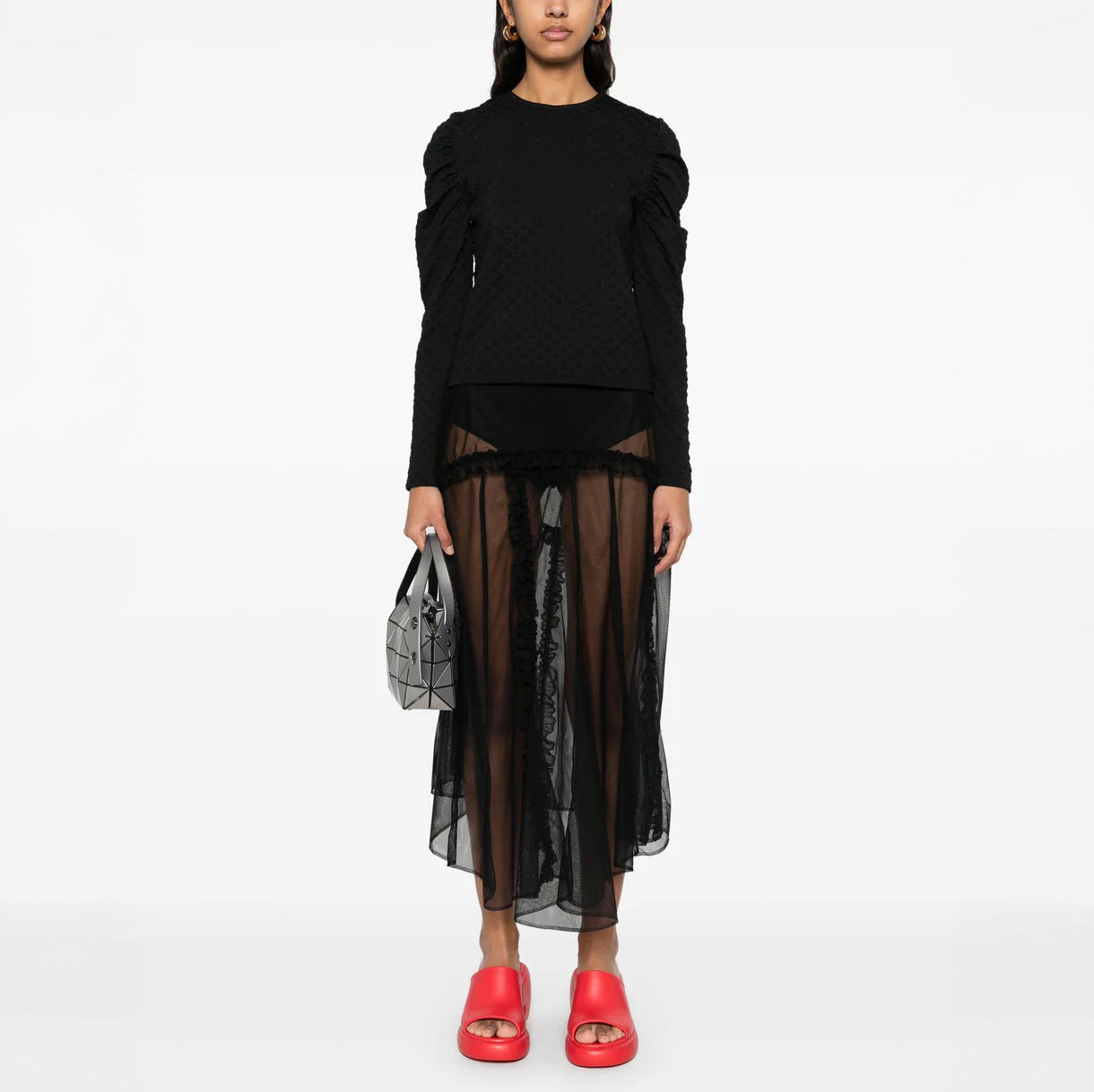 A woman stands facing forward wearing the Sun Reflection Blouse by Henrik Vibskov, paired with a sheer black skirt, bright red slide sandals, and holding a geometric silver handbag.